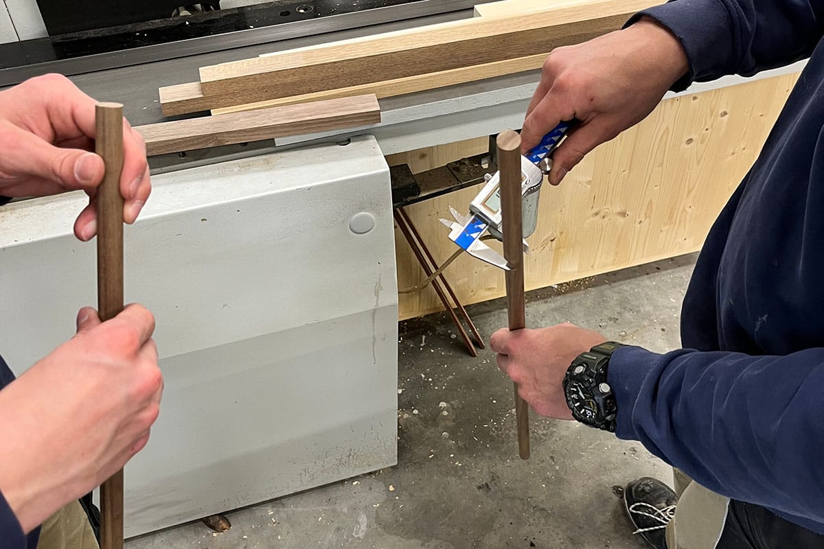 2025.03-FB-Frutiger-web-3 Zwei Personen halten jeweils ein rundes Holzstück in den Händen. Die Person auf der linken Seite hält das Holzstück mit beiden Händen, während die Person auf der rechten Seite das Holzstück mit einer Hand hält und in der anderen Hand ein Werkzeug hält. Im Hintergrund sind eine Werkbank, mehrere längliche Holzstücke und eine Holzwand zu sehen.